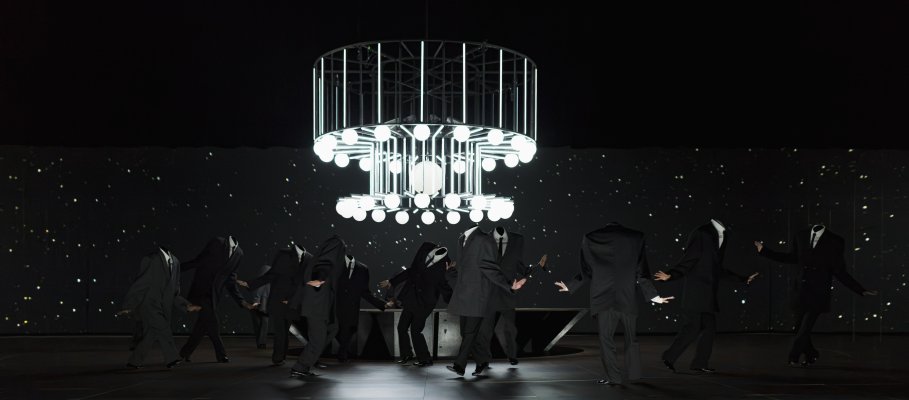 Several performers in dark suits move around a central stage platform. Costume elements conceal the heads, creating unusual silhouettes. Above them hangs a large lighting structure made of glowing spheres and metal frames.