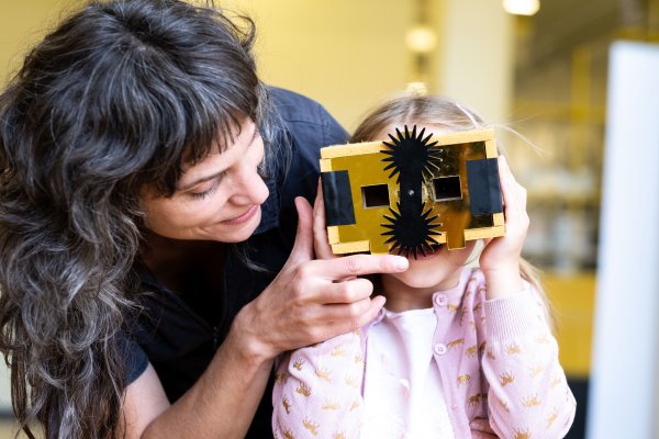 An adult supports a child using a handmade cardboard viewing device in a bright indoor space. The scene focuses on shared exploration, observation, and playful discovery through a creative object.