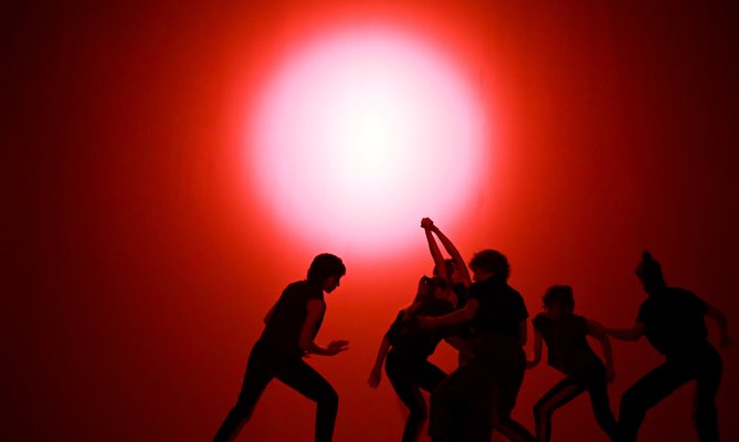 Panzetti, Ticcoi: All'Arme (c) Nina Durdevic Several performers move as silhouettes in front of an intensely red-lit stage. A large circular light dominates the background, framing the group and emphasizing overlapping body shapes and gestures.