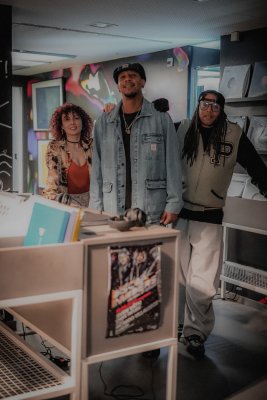 Drei Personen stehen nebeneinander in einem Raum mit Schallplattenregalen und Ausstellungsm&ouml;beln. Medien, Kopfh&ouml;rer und Wandgrafiken bilden den Rahmen f&uuml;r die Gruppenaufnahme.