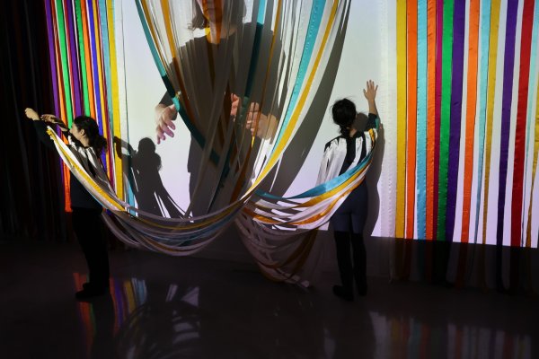 Zwei Personen stehen vor einer wandf&uuml;llenden Installation aus farbigen Stoffb&auml;ndern. Einige B&auml;nder h&auml;ngen vertikal, andere bilden B&ouml;gen im Raum. Projektionen und Schatten &uuml;berlagern die Szene und erzeugen zus&auml;tzliche Ebenen.