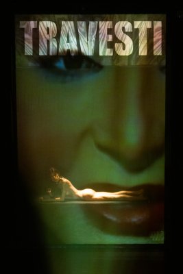 Renata Carvalho, MANIFESTO TRANSPROFÁGICO Das Foto zeigt eine einzelne Performer*in auf einer Bühne, die auf dem Bauch vor einer großen Projektionsfläche liegt. Über der Person erscheint das projizierte Wort „TRAVESTI“ in großen Buchstaben.