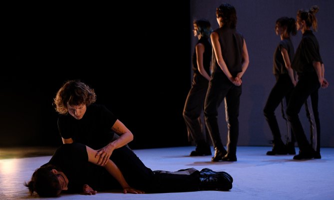 Panzetti, Ticcoi: All'Arme (c) Nina Durdevic On a bright stage floor, one performer lies on the ground while another leans over them. Several performers stand in the background. The restrained color palette draws attention to bodily positions and spatial relationships.