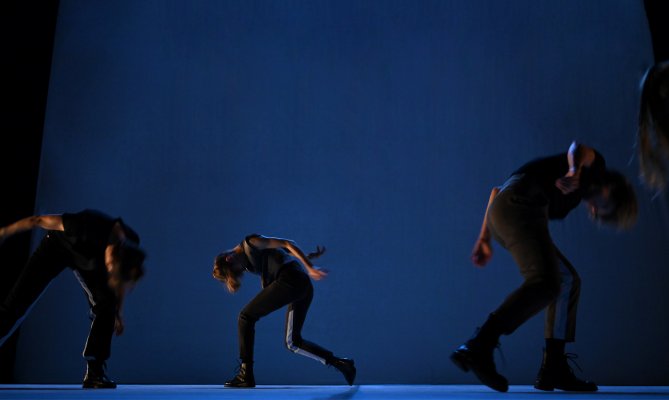 Panzetti, Ticcoi: All'Arme (c) Nina Durdevic Three performers move in front of a large blue-lit backdrop. Their bodies lean in different directions, creating dynamic lines through arms, legs, and torsos. The lighting emphasizes movement, spacing, and the overall stage composition.