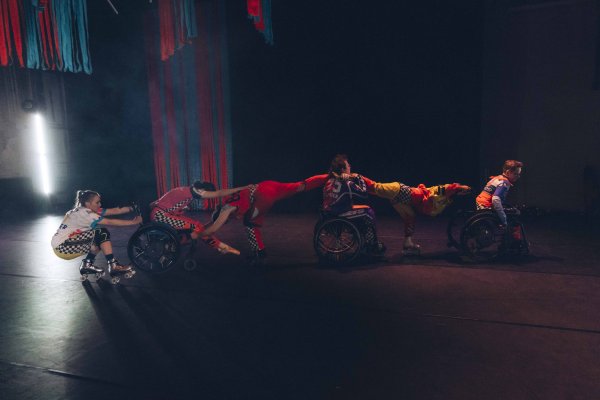 Eng Kai Er: The Wheelie Show (c) Alexandra Polina Mehrere Performende mit Rollschuhen und Rollstühlen sind auf einer dunklen Bühne zu einer waagerechten Bewegungssequenz verbunden. Bunte Kostüme und gerichtetes Licht betonen die gemeinsame Formation sowie das Zusammenspiel von Körpern und Rädern.