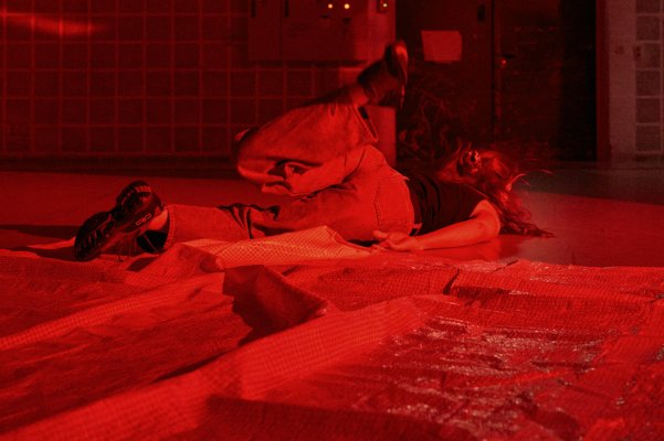 Eine Person liegt auf dem Bauch am Boden einer B&uuml;hne. Das linke Bein hebt sich nach oben und erzeugt den Eindruck von Bewegung. Intensives rotes Licht taucht die gesamte Szene in einen monochromen Farbton.